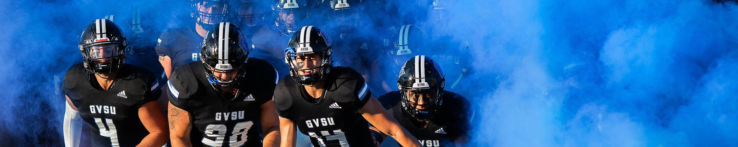 Football players in black and white uniforms run onto the field through a cloud of blue smoke.